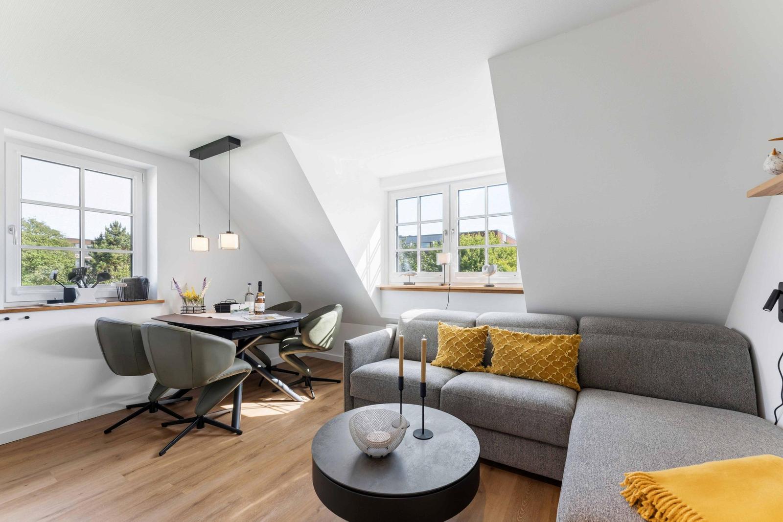 Wohn- und Essbereich mit Sofa, Tisch und Stühlen. Holzboden und große Fenster.