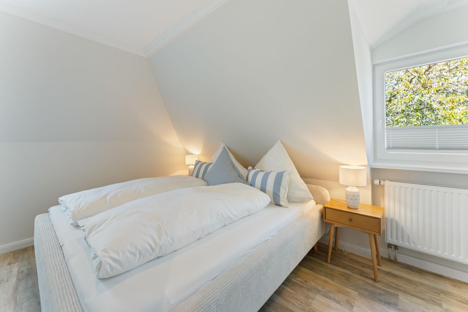 Schlafzimmer mit Doppelbett, Nachttisch und Fenster mit Blick auf Bäume.
