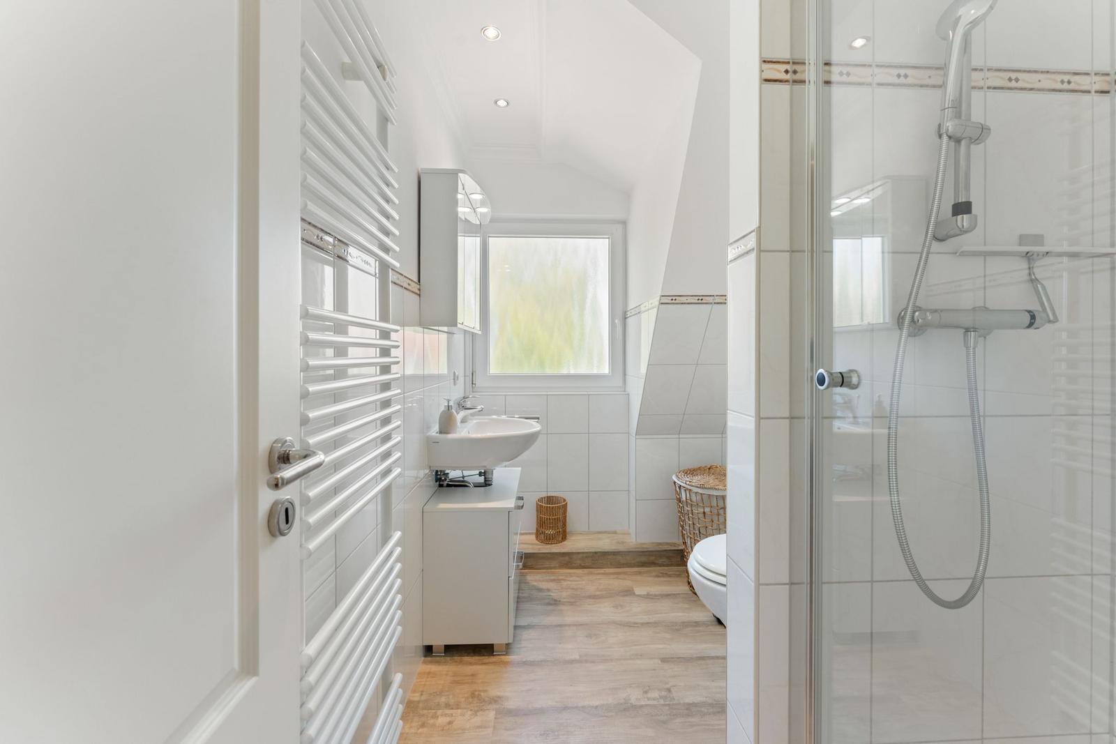 Badezimmer mit Dusche, Waschbecken und Toilette. Holzfußboden und Fenster.
