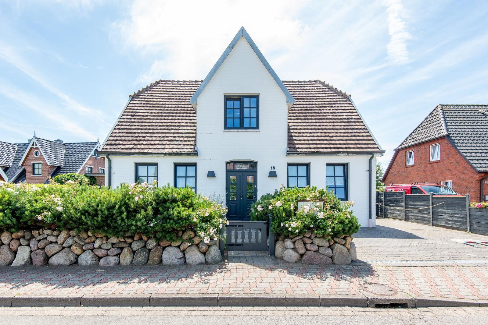 Weiße Villa mit Steinmauer und Blumenbeeten. Schwarze Fenster und Tür. Zufahrt mit Pflastersteinen.