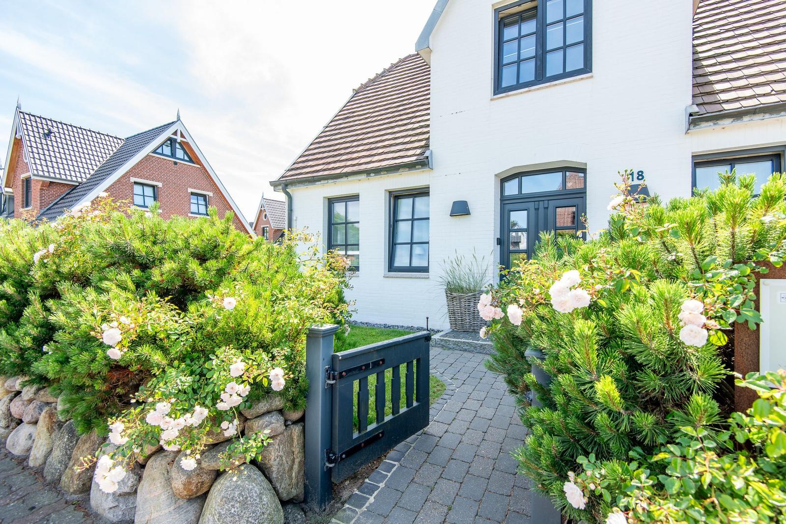 Haus mit weißer Fassade, dunklen Fenstern und gepflastertem Weg durch grüne Hecken.