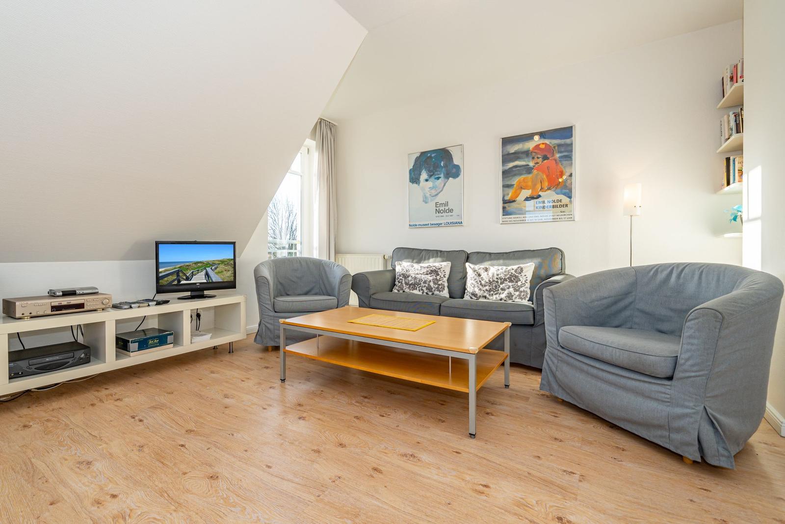 Wohnzimmer mit Sofa, zwei Sesseln, Couchtisch und Fernseher auf Regal.