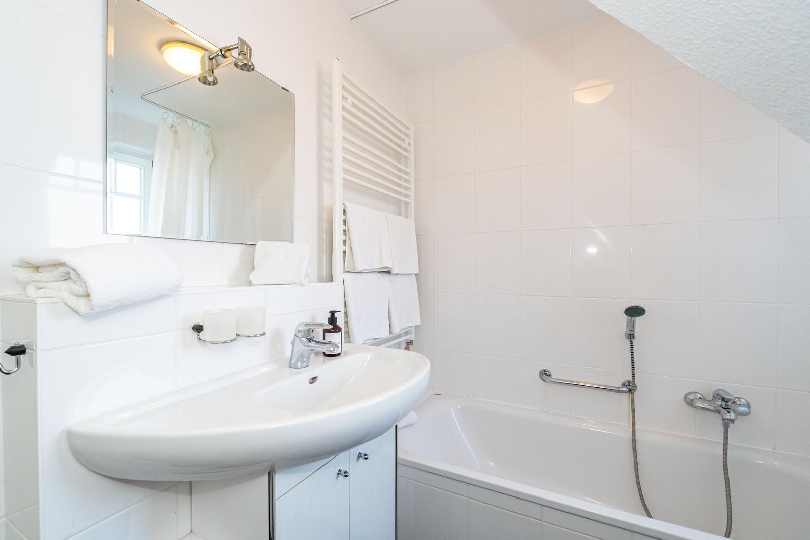 Badezimmer mit Badewanne, Waschbecken und Spiegel. Weiße Fliesen und Handtücher.