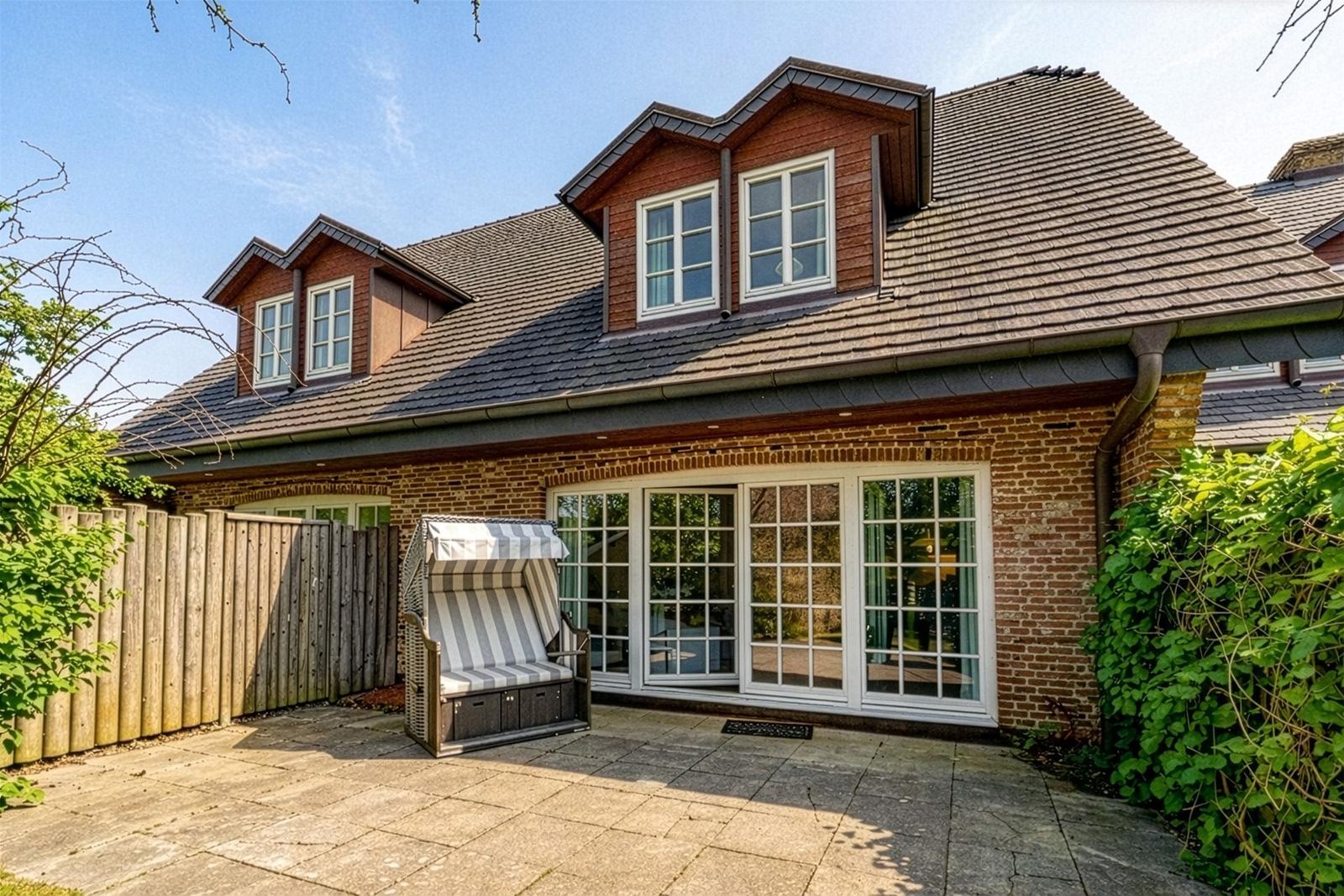 Haus mit Holzschindeldach, großen Fenstern und Terrasse mit Liegestuhl.