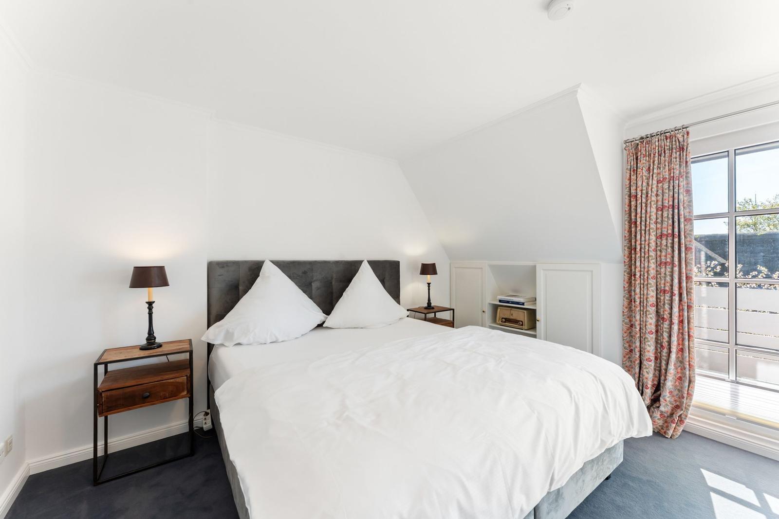 Schlafzimmer mit Doppelbett, Nachttisch und Fenster mit Vorhängen.