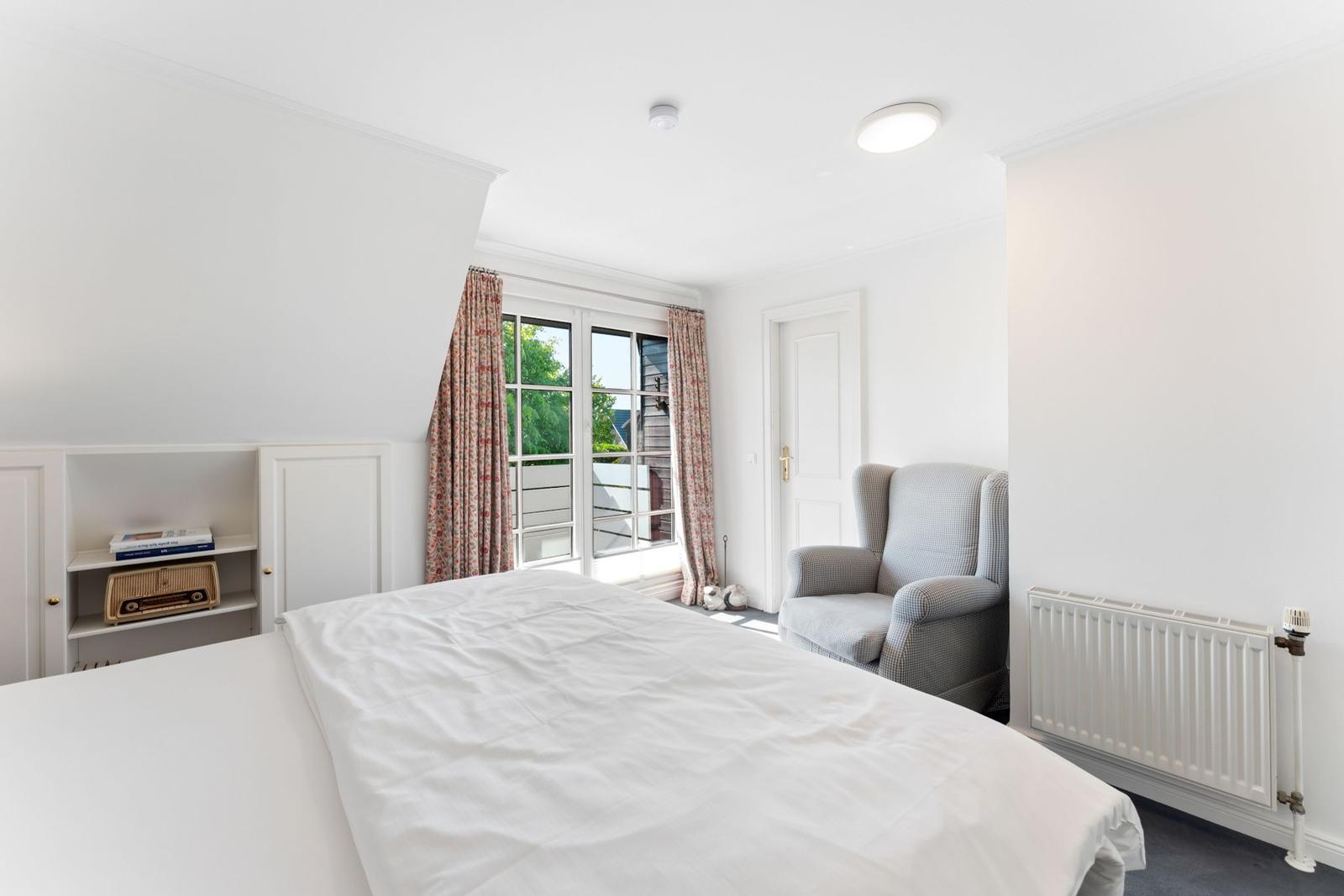 Schlafzimmer mit Bett, Sessel, Fenster und Schrank. Vorhang und Heizkörper sind sichtbar.