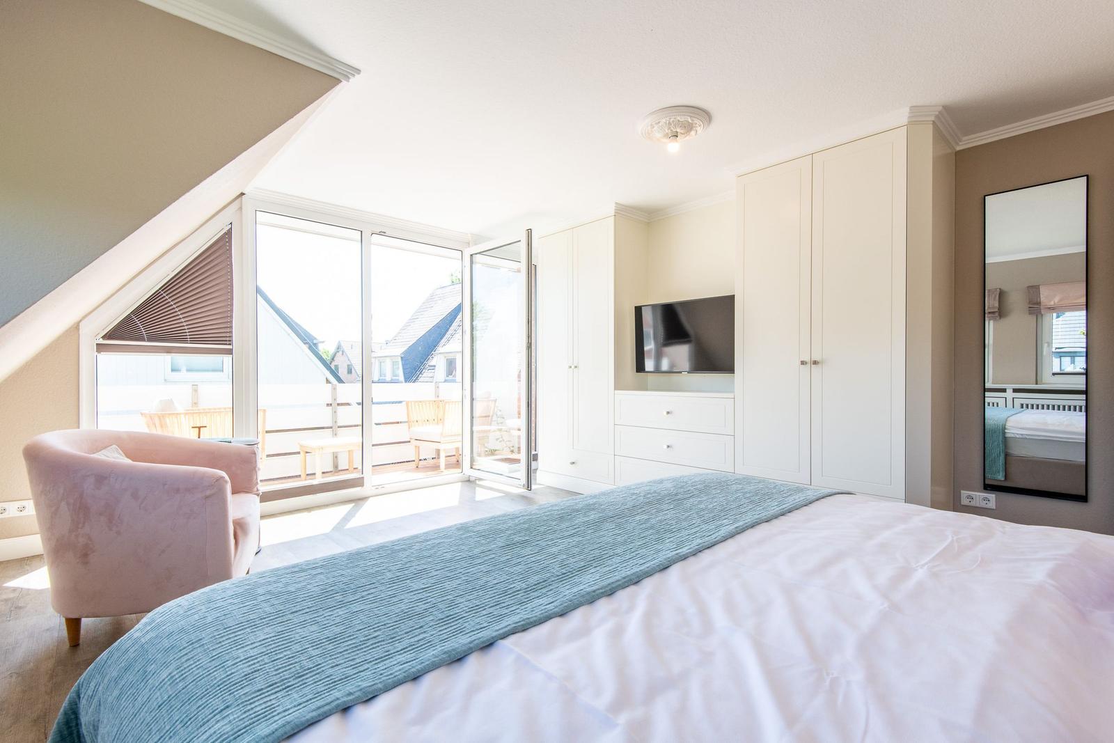 Schlafzimmer mit Bett, Schrank und TV. Balkon über großes Fenster. Rosa Sessel.