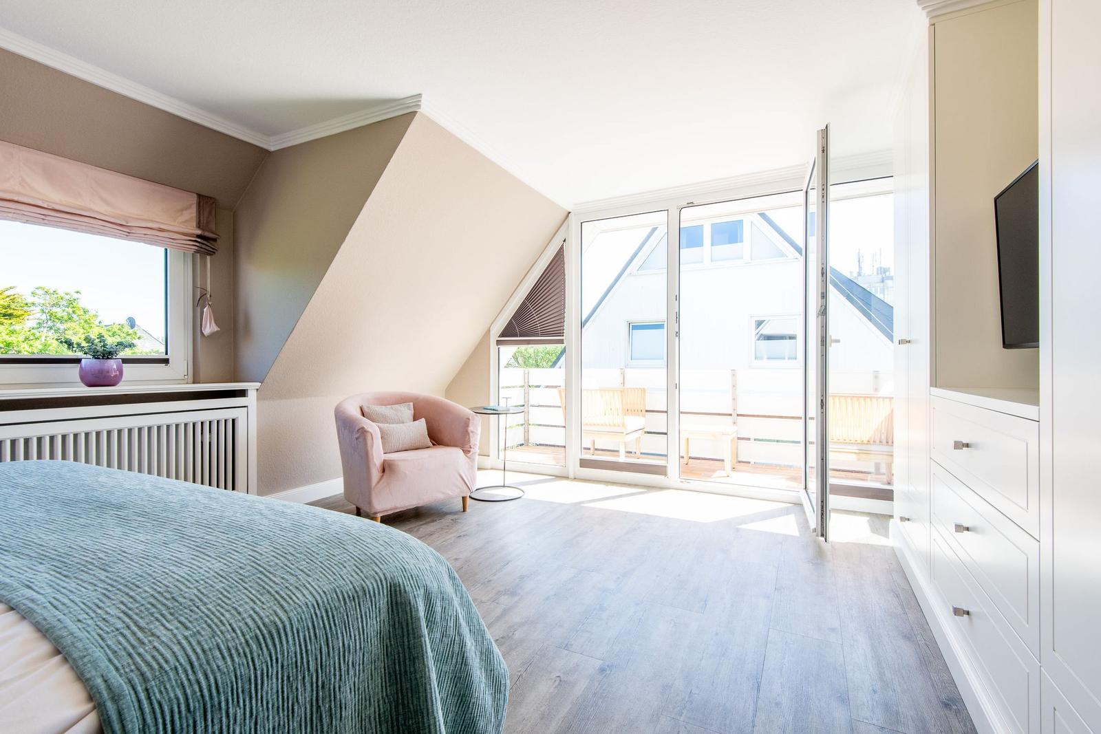 Schlafzimmer mit Balkon, Bett, Sessel und Schrank. Fenster und Tür sind geöffnet.