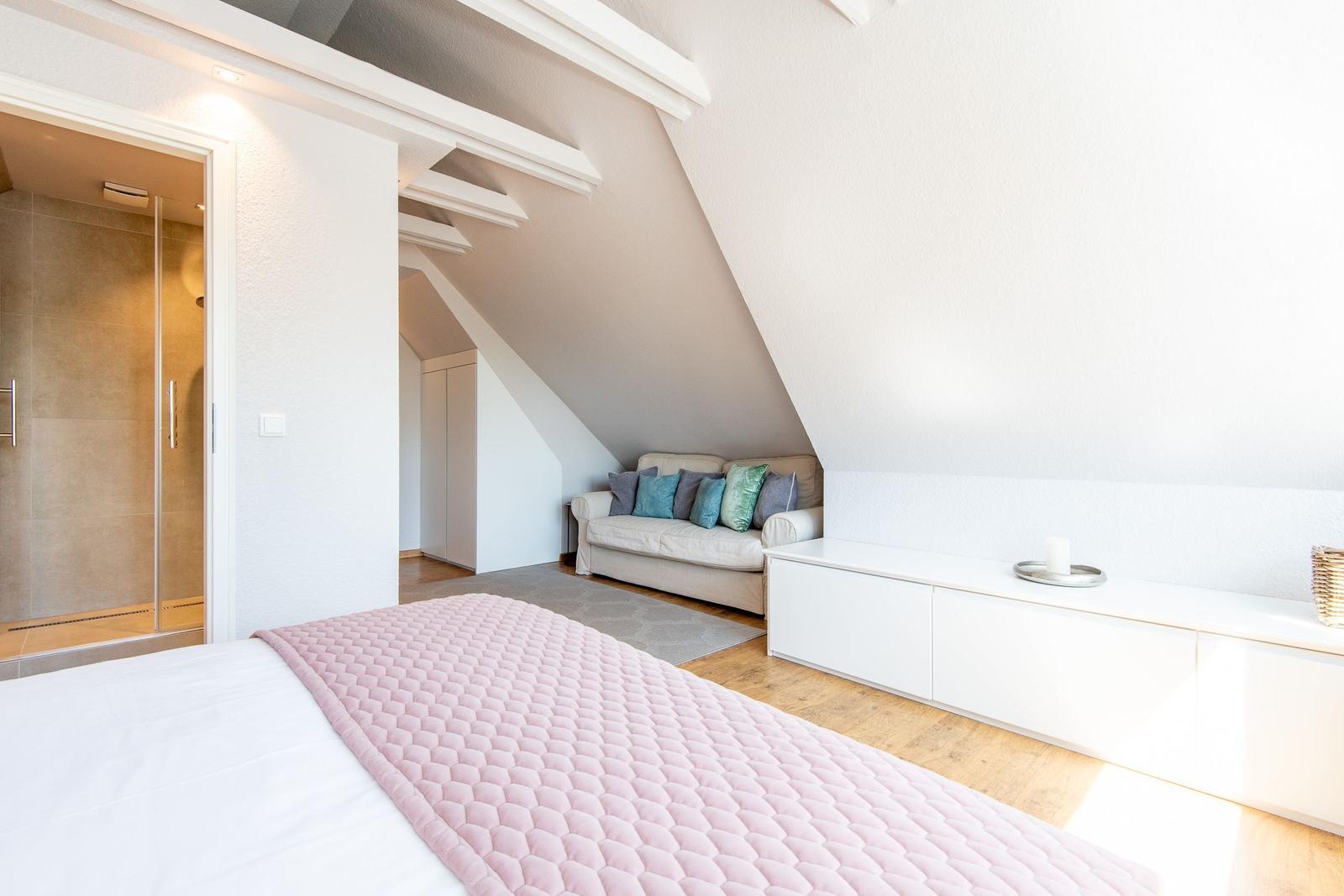 Schlafzimmer mit Dachschräge, Sofa und Bad. Bett mit rosa Decke.