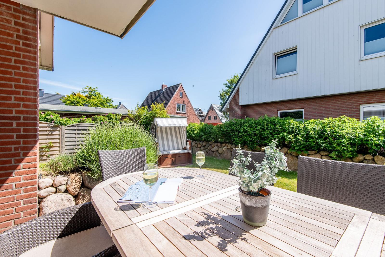 Terrasse mit Tisch, Stühlen und Sonnenschirm. Hintergrund: Garten, Hecke und Nachbarhäuser.