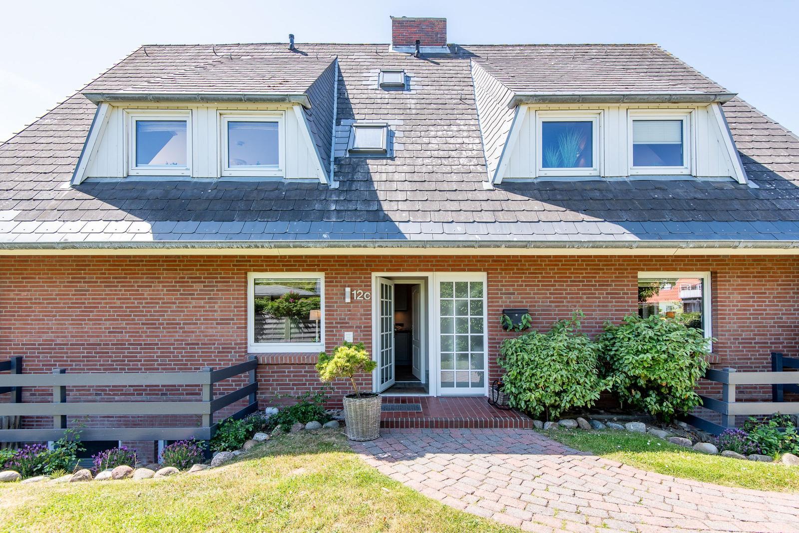 Haus mit rotem Backsteinfassade, Dachgauben und gepflasterter Eingangsbereich mit Pflanzen.