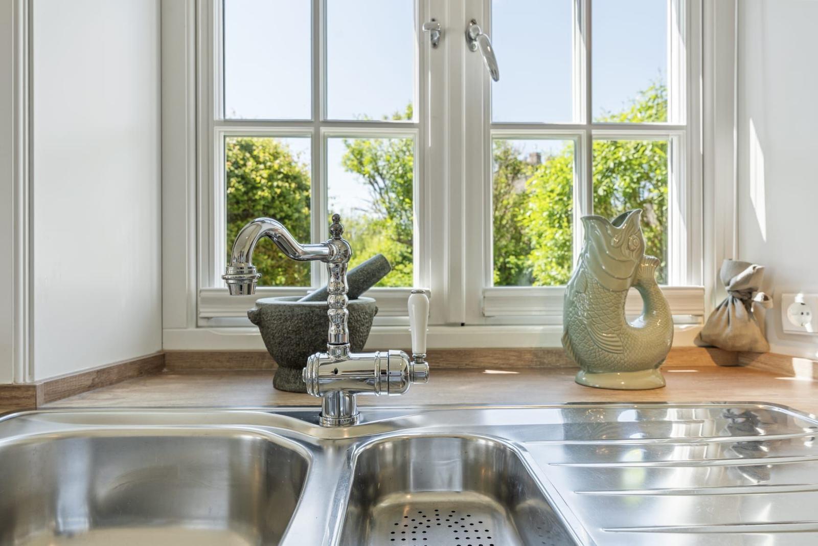 Küchenarm mit Edelstahlwaschbecken, Fenster mit Blick auf Grün, Fischfigur und Mörser auf Arbeitsfläche.