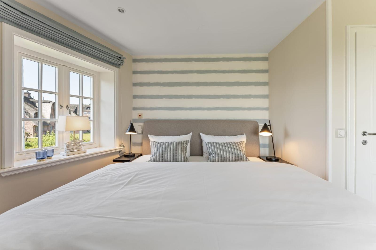 Schlafzimmer mit großem Bett, Fenster mit Blick und gestreifter Wand hinter dem Kopfteil.