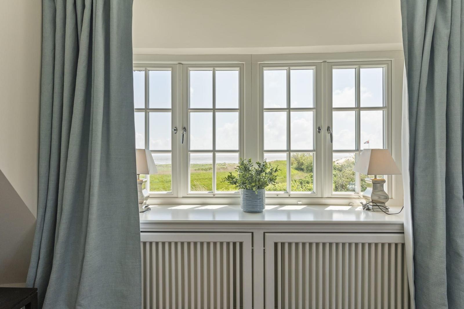 Grosses Fenster mit Blick auf Grünland und Meer. Auf Fensterbank stehen zwei Lampen und eine Pflanze.