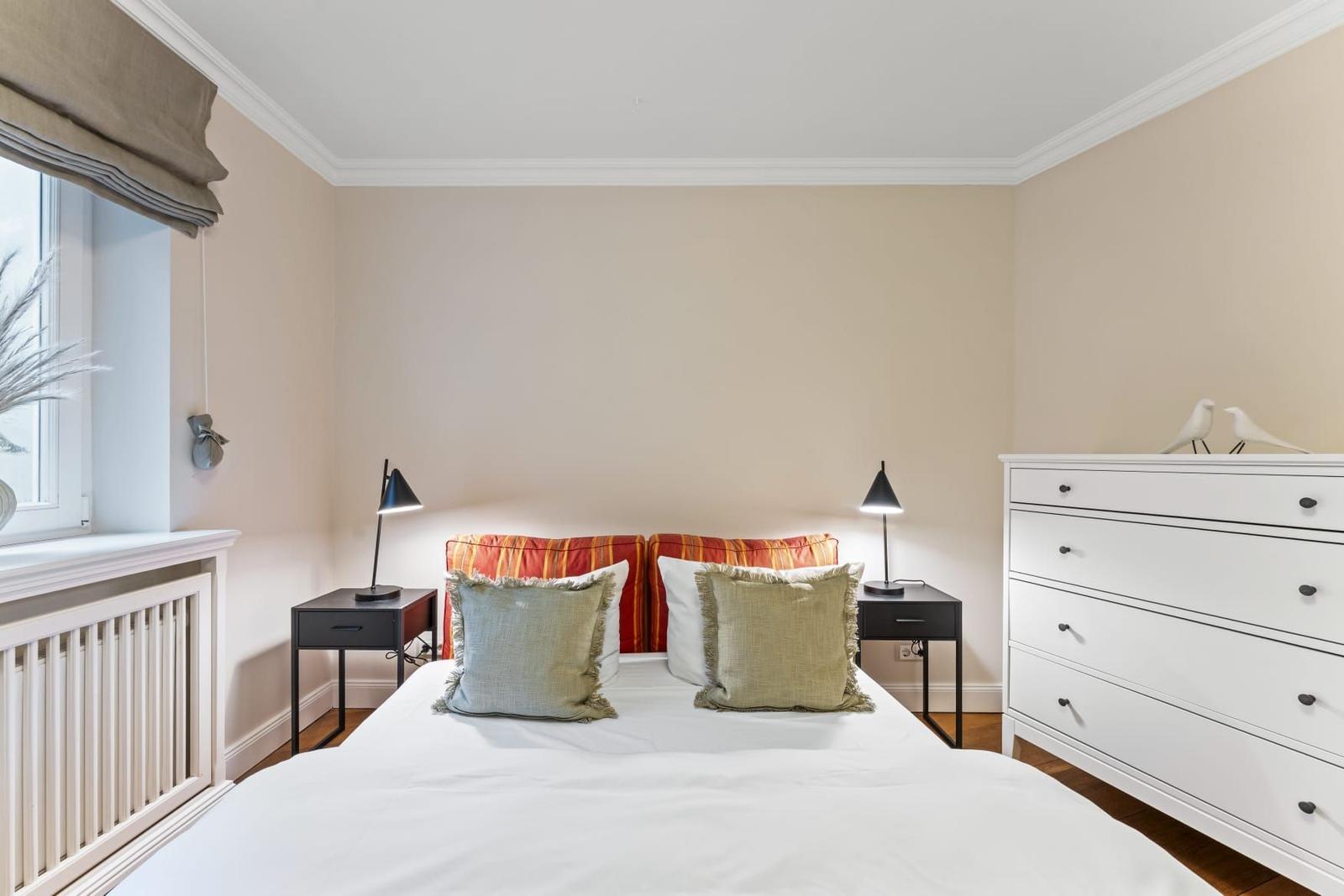 Schlafzimmer mit Bett, Nachttischen und Kleiderschrank. Fenster mit Rollladen und dekorativen Kissen.