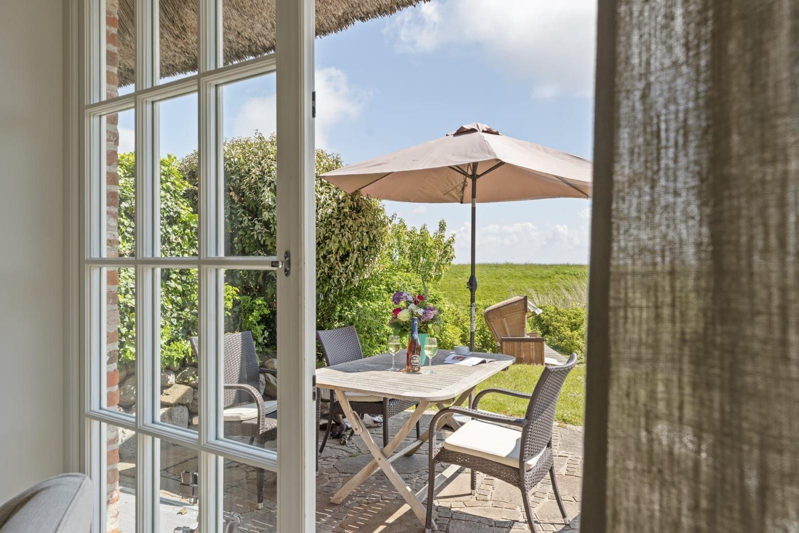 Durch die Glastür blickt man auf eine Terrasse mit Tisch, Stühlen und Sonnenschirm.
