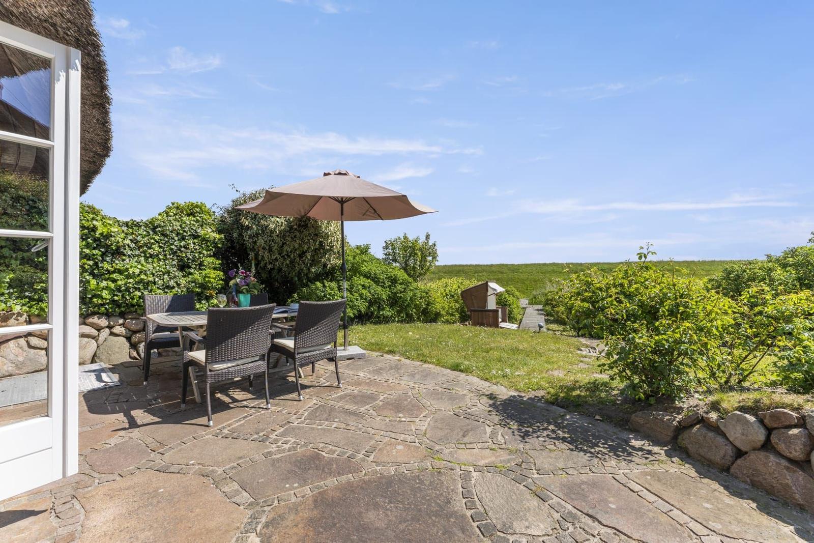Terrasse mit Tisch, Stühlen und Sonnenschirm. Steinplatten, Gras und Sträucher im Hintergrund.