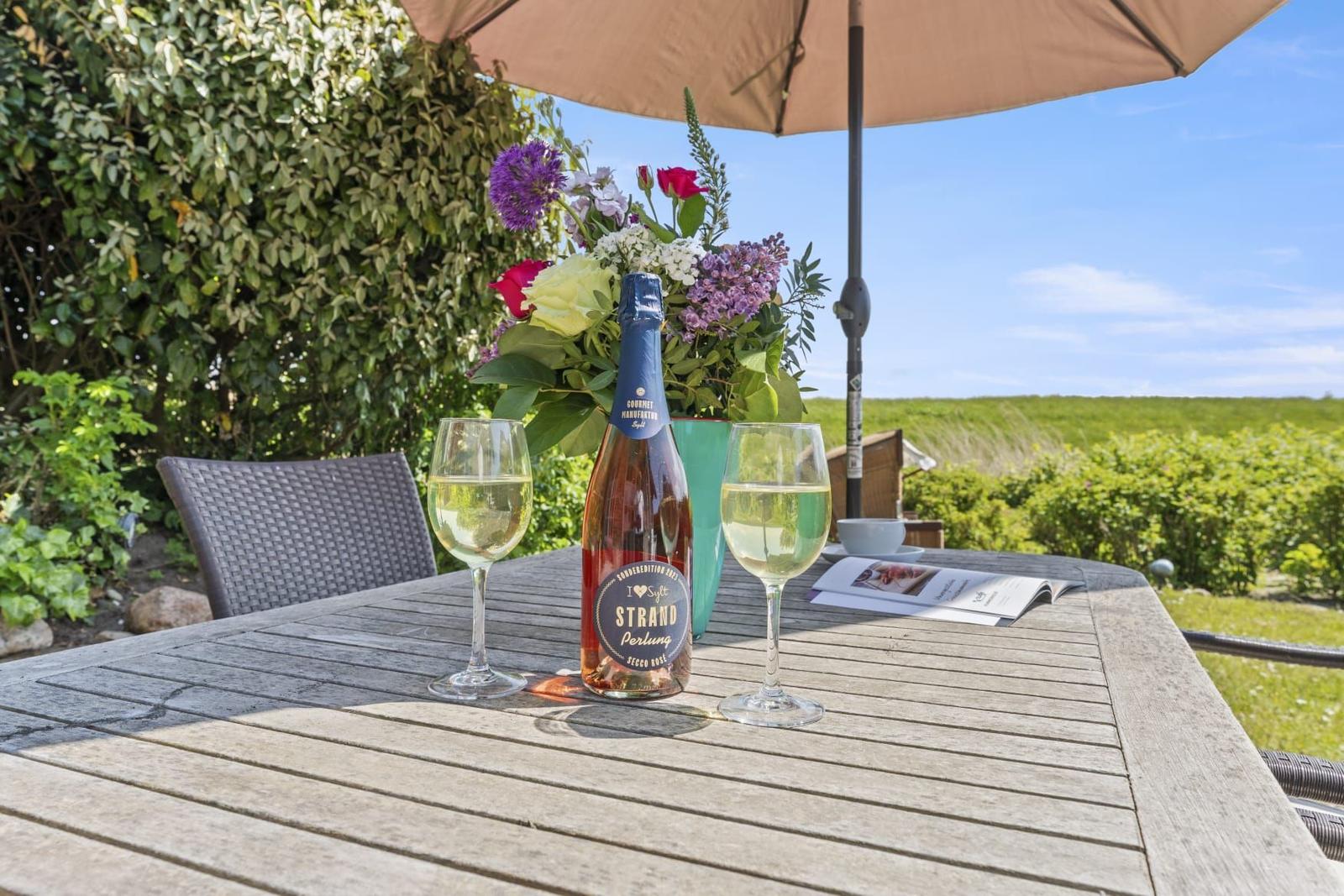 Terrasse mit Tisch, Stühlen, Blumen und Flasche Rosé unter Sonnenschutz.