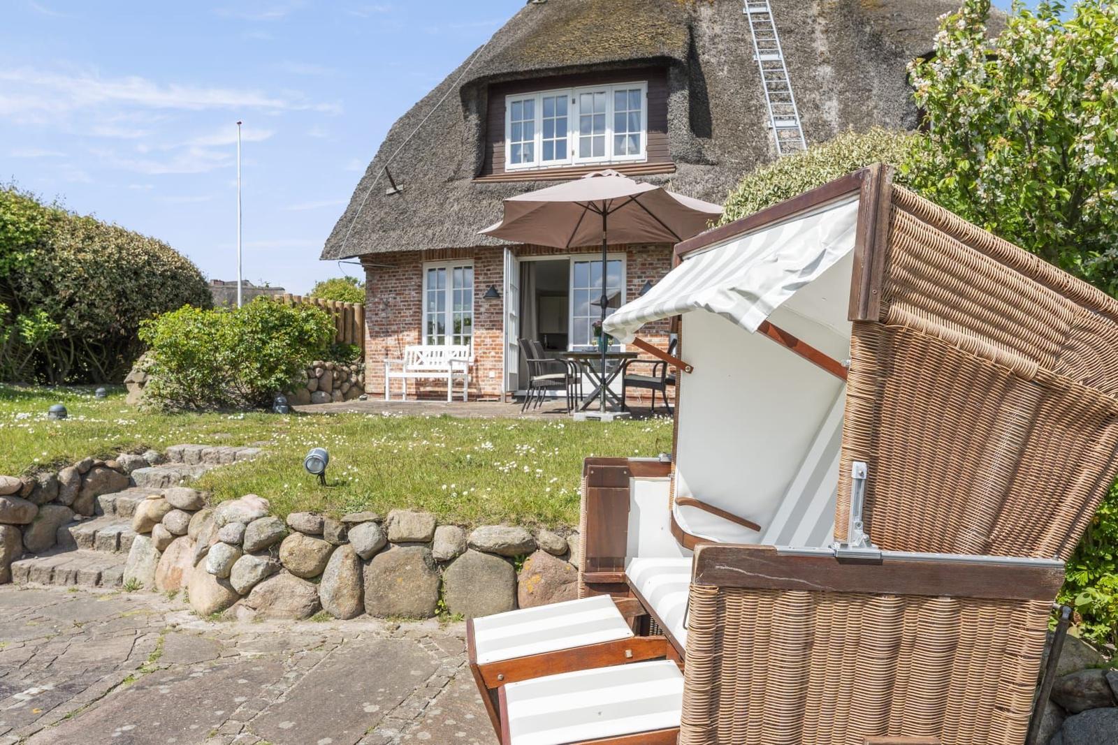 Haus mit Strohdach, Gartenbereich mit Liegestuhl und Terrasse mit Tisch und Stühlen.