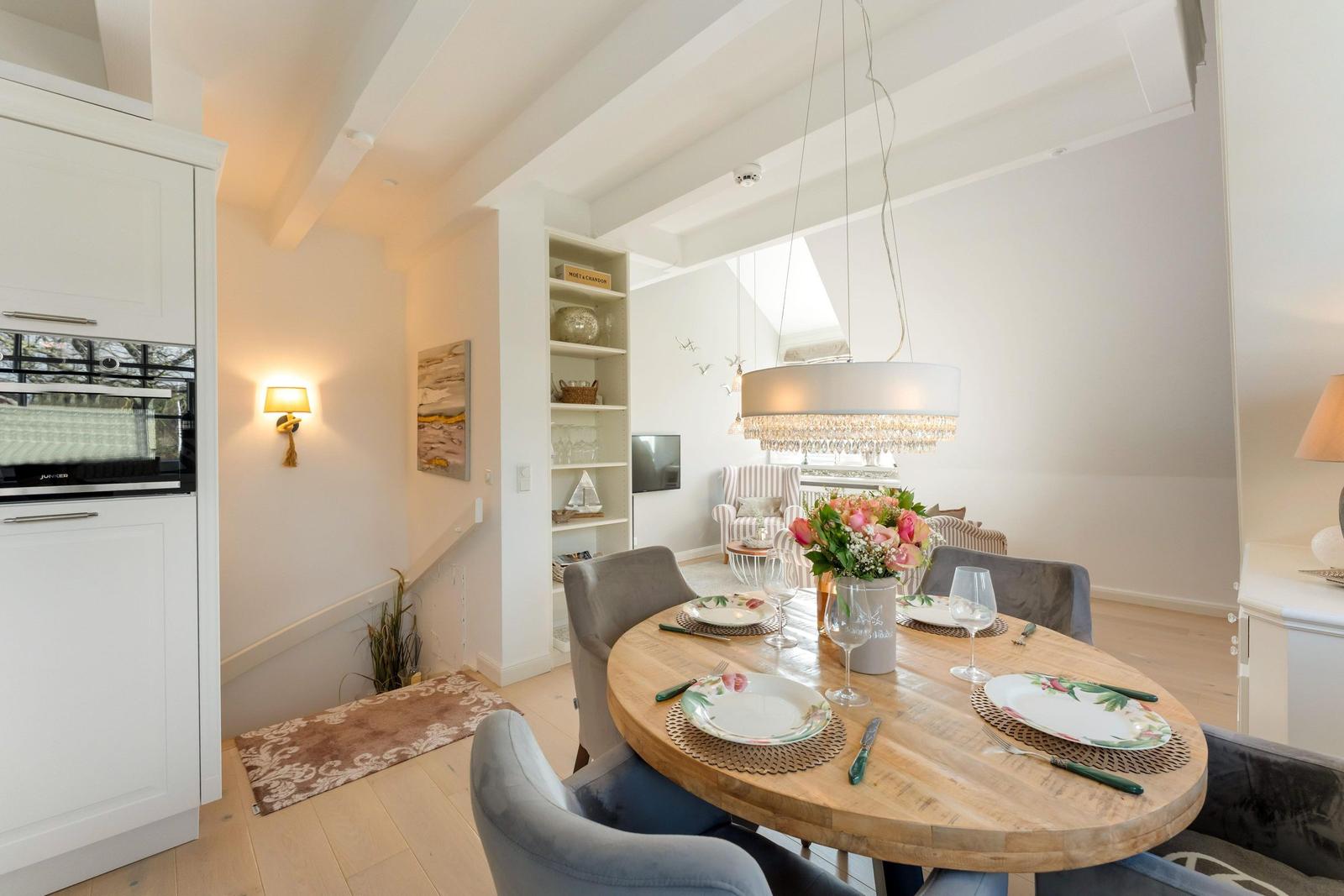 Dining area with round table, gray chairs, and floral decoration. Kitchen and living area are visible.