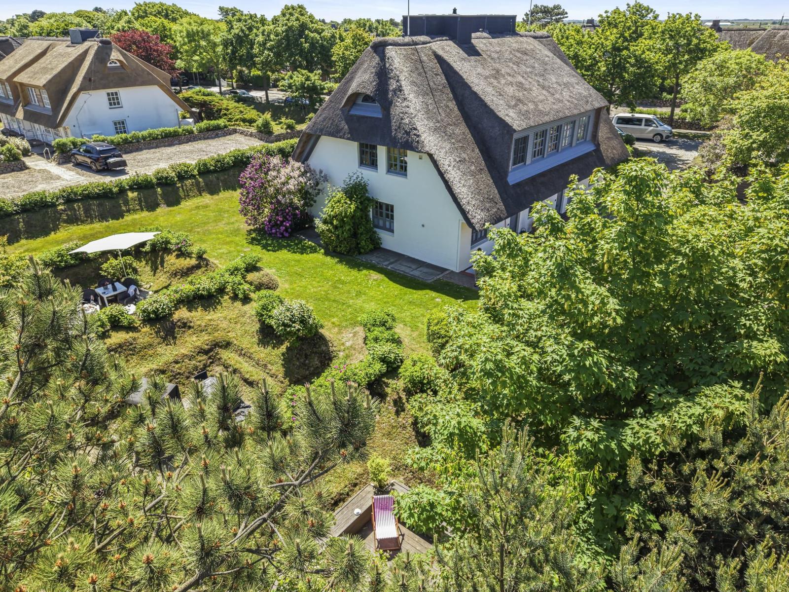 House with thatched roof, garden with umbrella and lounge chairs, surrounded by trees.