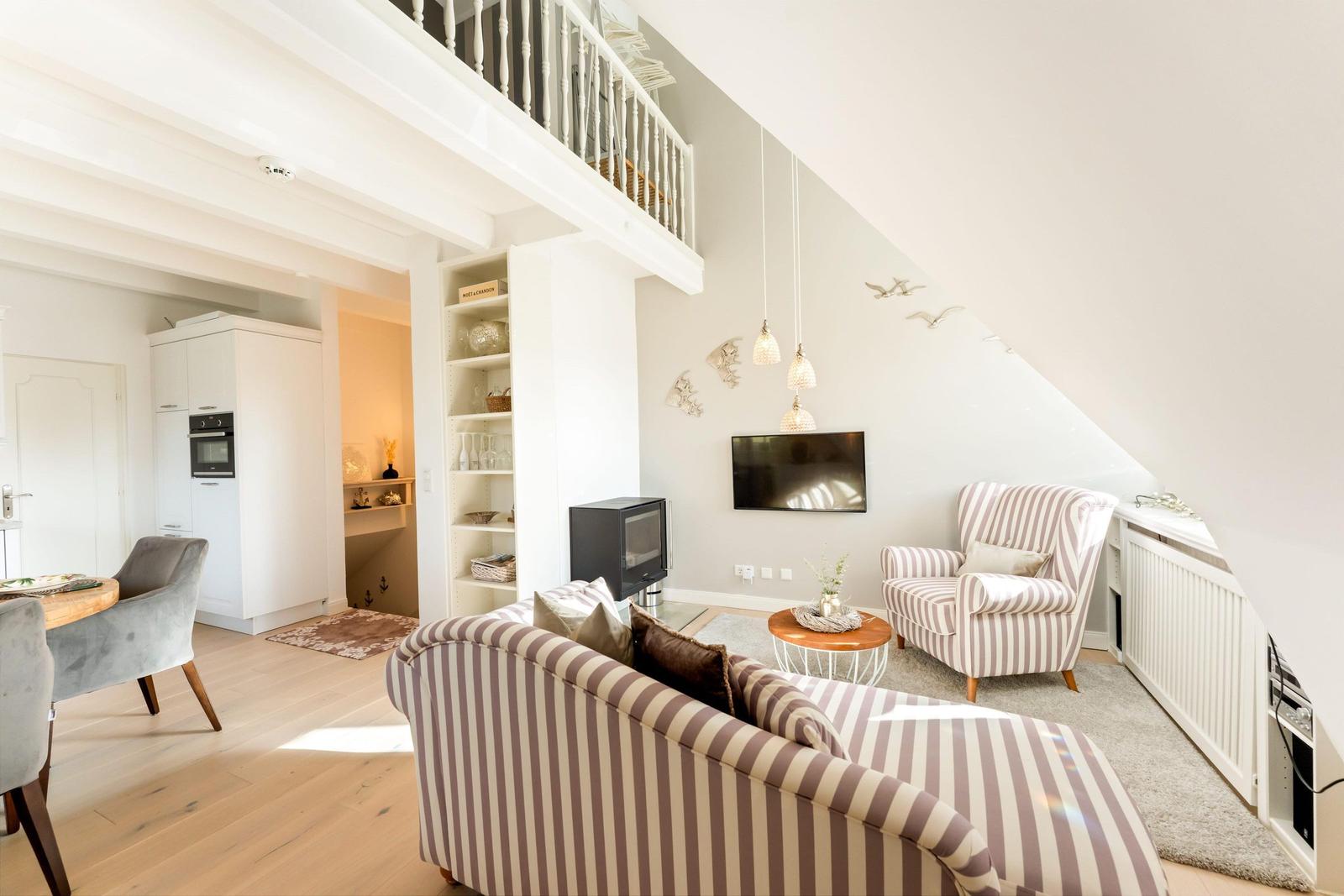 Living room with sofa, armchair, and TV under sloped ceilings. Kitchen and dining area are visible.