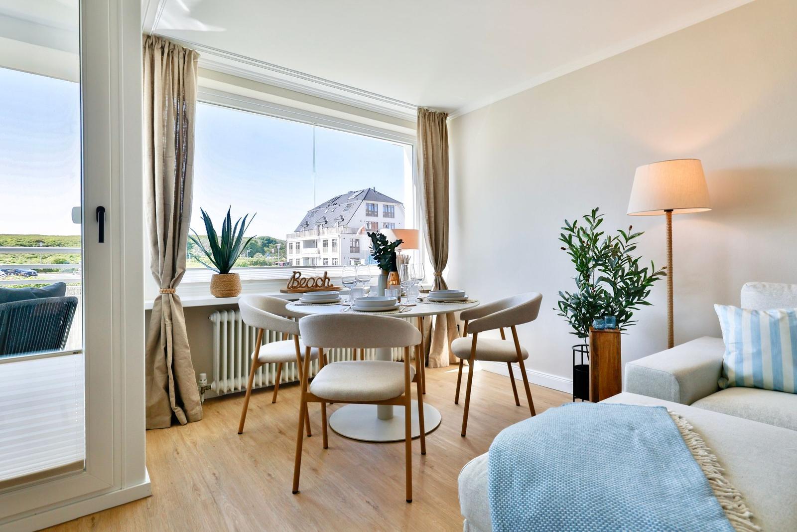 Essbereich mit Tisch und Stühlen, Blick auf Gebäude durch Fenster. Sofa mit Kissen und Beistelltisch.