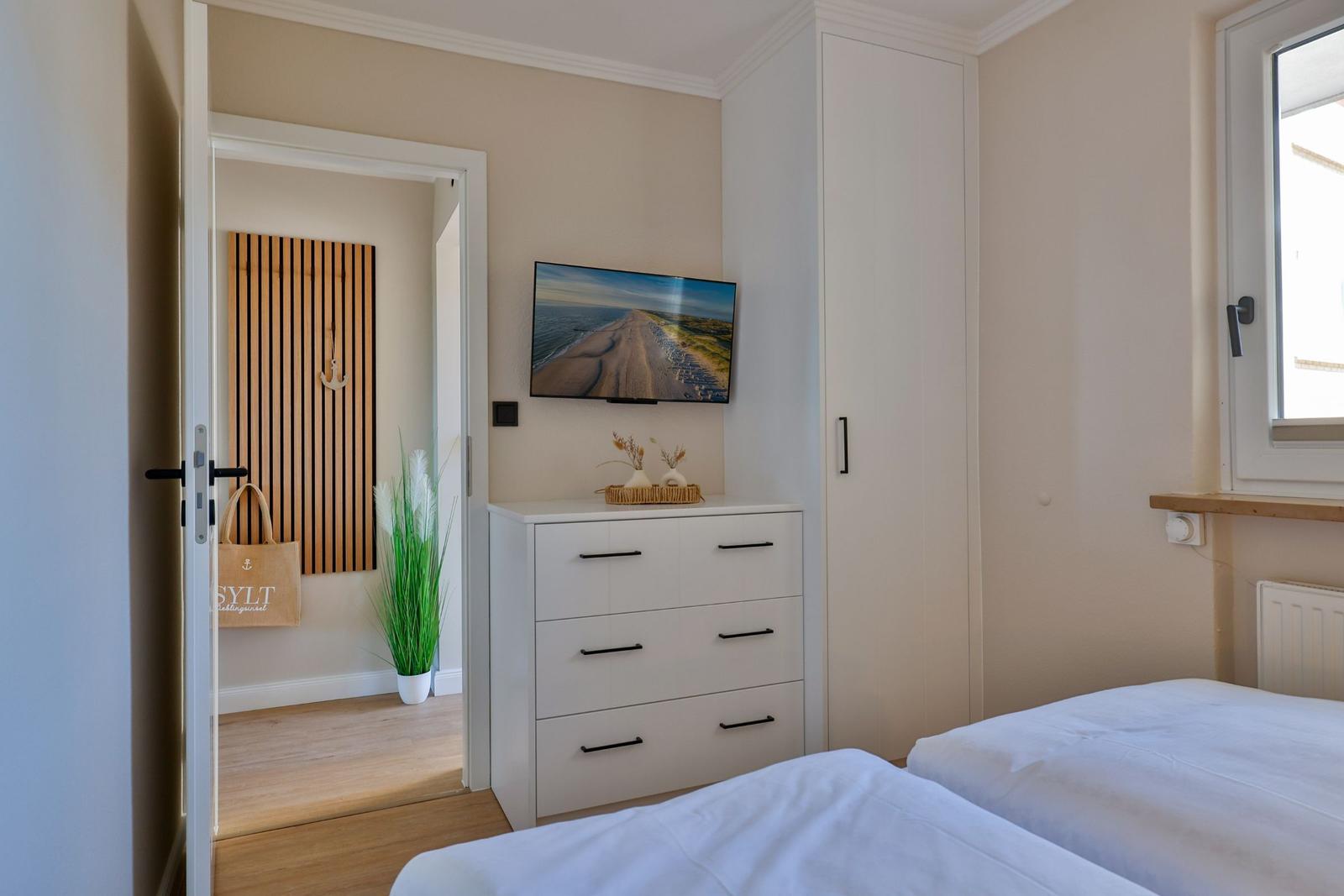 Schlafzimmer mit zwei Betten, Schrank und Fernseher. Tür führt in einen Flur mit Holzpaneelen.