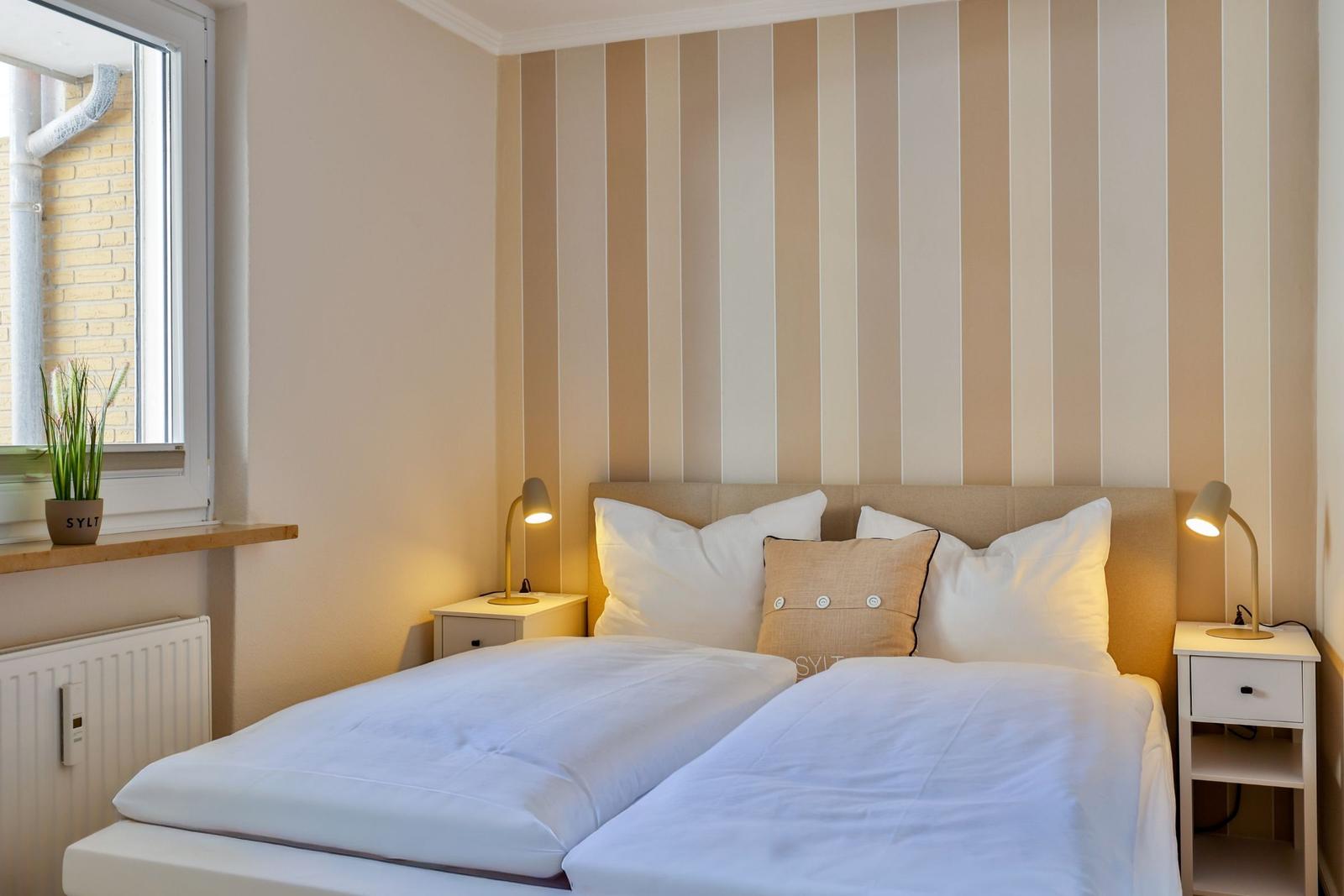 Schlafzimmer mit Bett, zwei Nachttischen und Streifenwand. Fenster mit Pflanze und Heizung.