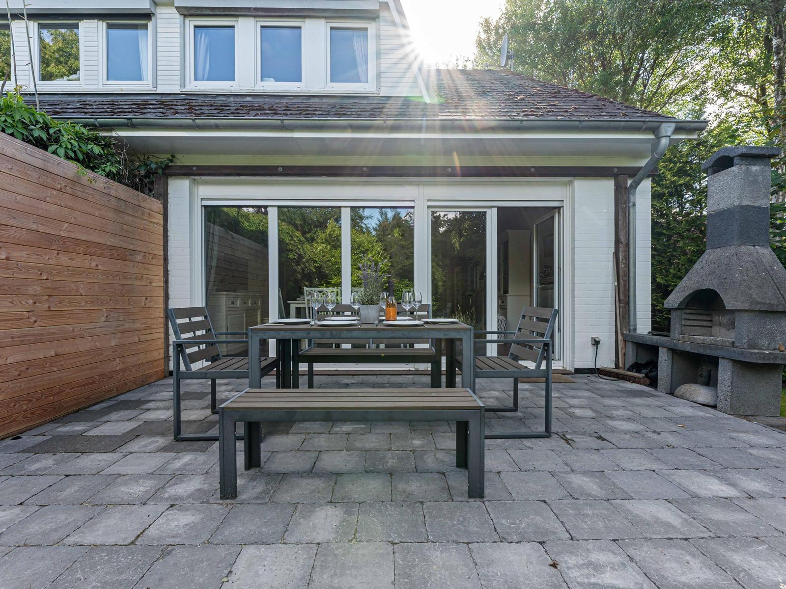 Terrasse mit Tisch, Bänken und Grillofen. Hintergrund: Haus mit Glasfront und Holzmauer.