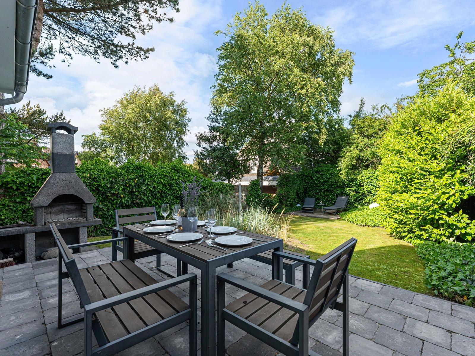 Terrasse mit Tisch, Stühlen und Grill. Grünflächen und Bäume im Hintergrund.