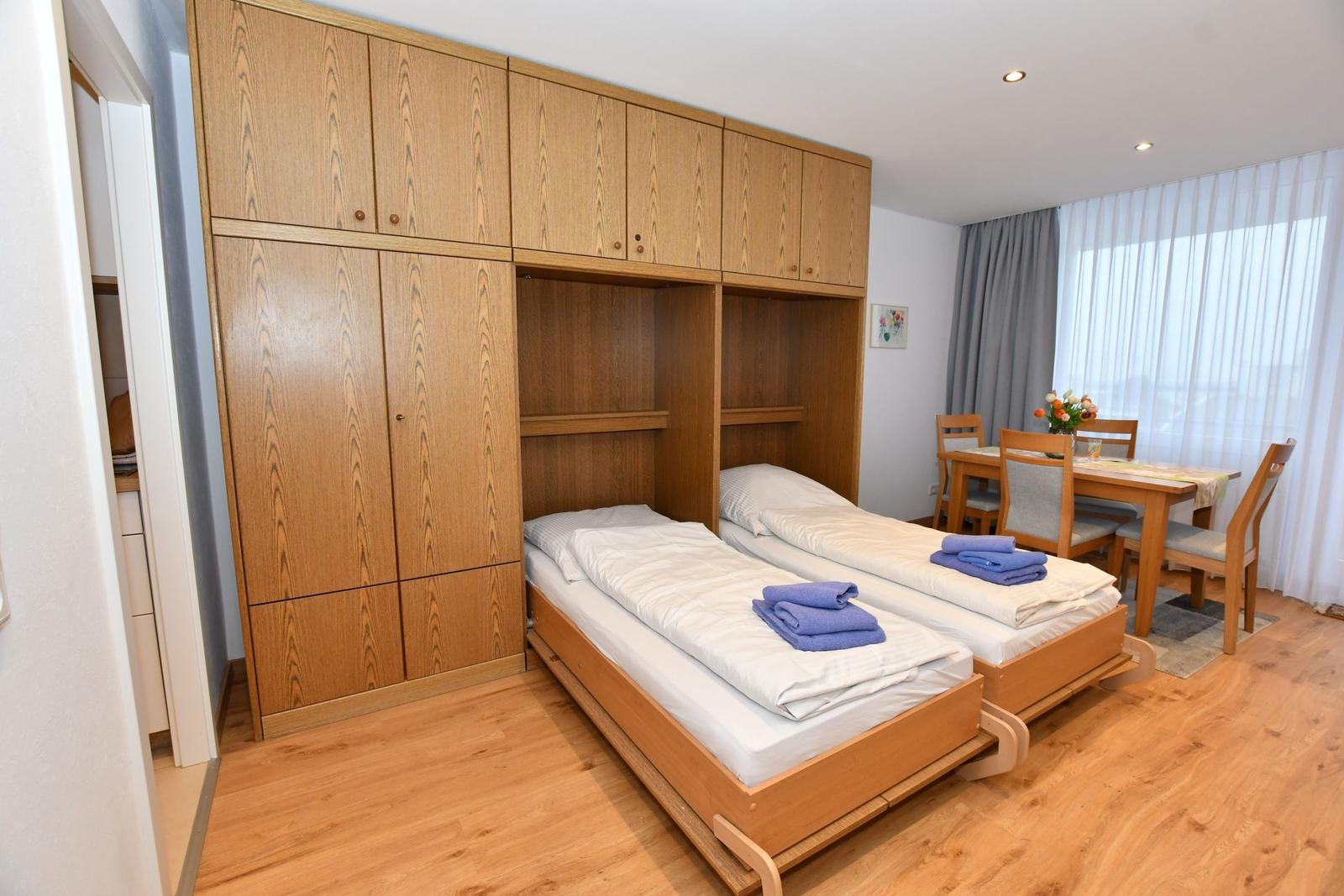Zwei Einzelbetten mit Laken und Handtüchern. Holzmöbel und Essbereich im Hintergrund.