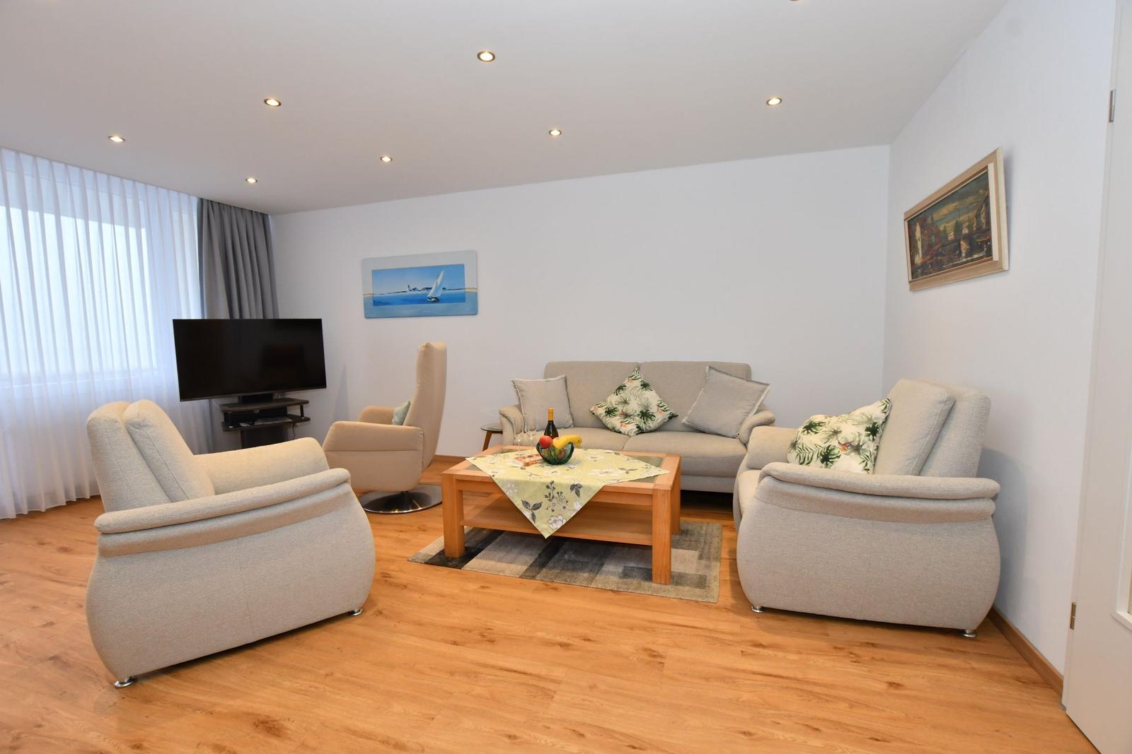 Wohnzimmer mit Sofa, Sesseln, Couchtisch und Fernseher. Holzboden und Wandbilder.