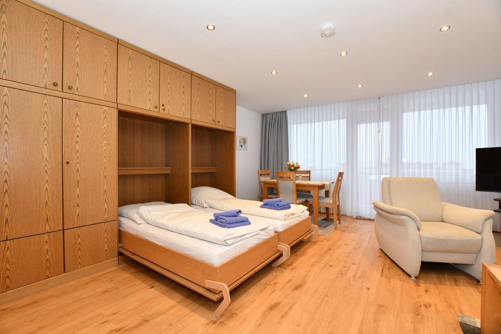Zimmer mit Schlafcabinen, Esstisch und Sessel. Holzboden und große Fenster.
