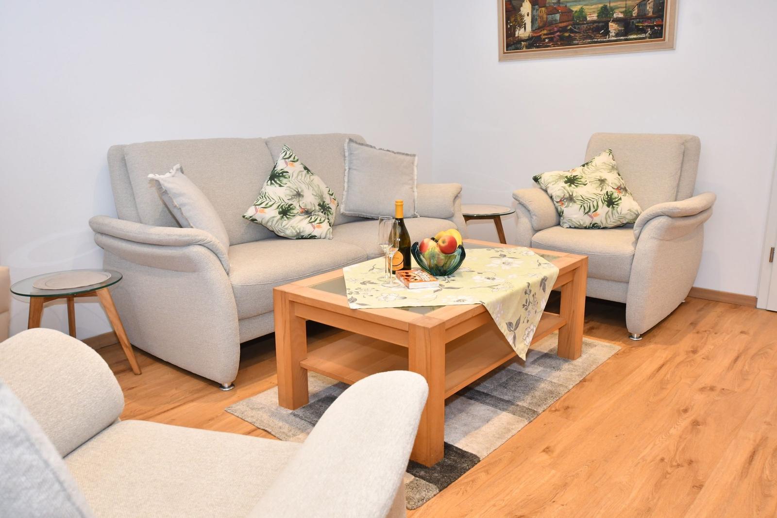 Wohnzimmer mit Sofa, Sessel und Holztisch. Auf dem Tisch steht eine Flasche Wein und Obst.