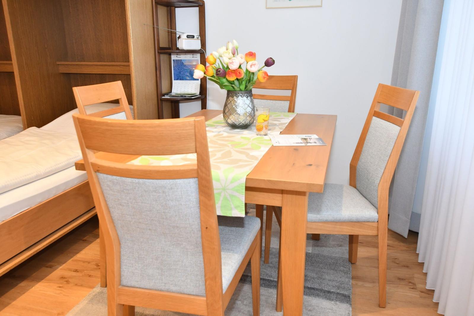 Esstisch mit vier Stühlen, Blumen und Tischdecke in einem Raum mit Bett und Holzmöbeln.