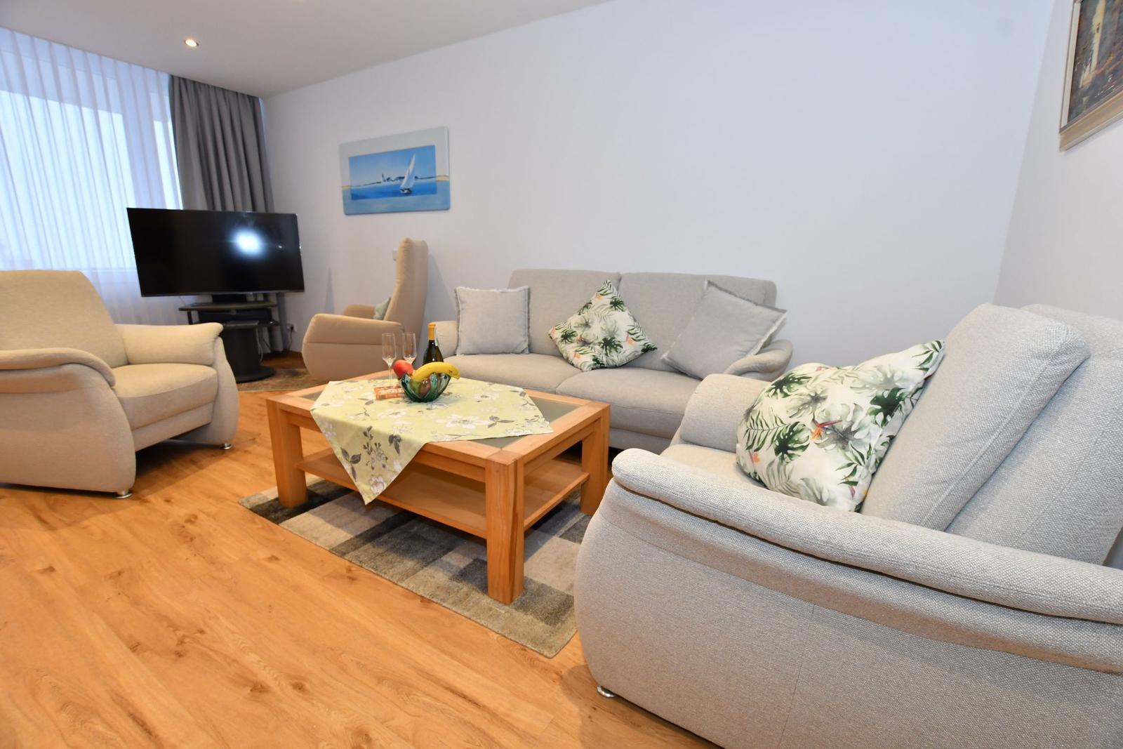 Wohnzimmer mit Sofa, Sesseln und Holztisch. Fernseher und Wandbild sind sichtbar.
