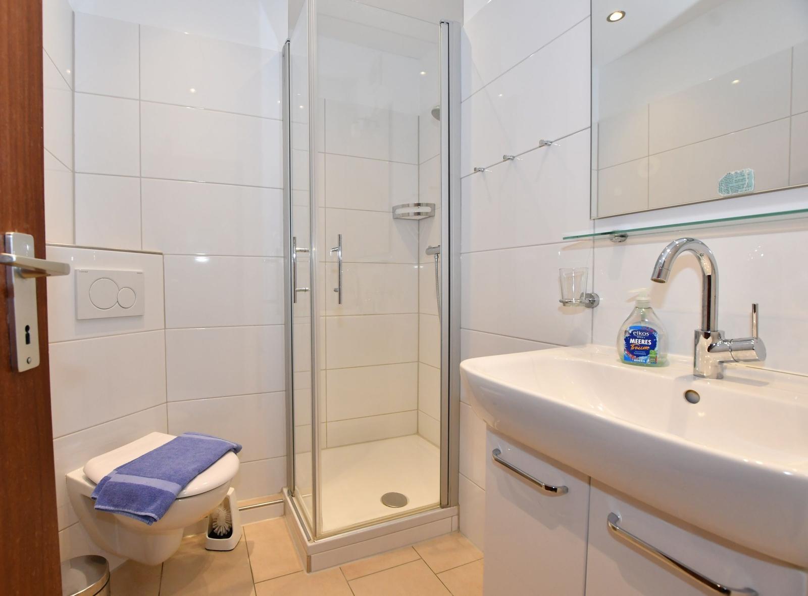 Badezimmer mit Dusche, Waschbecken und Toilette. Weiße Fliesen und moderne Einrichtung.