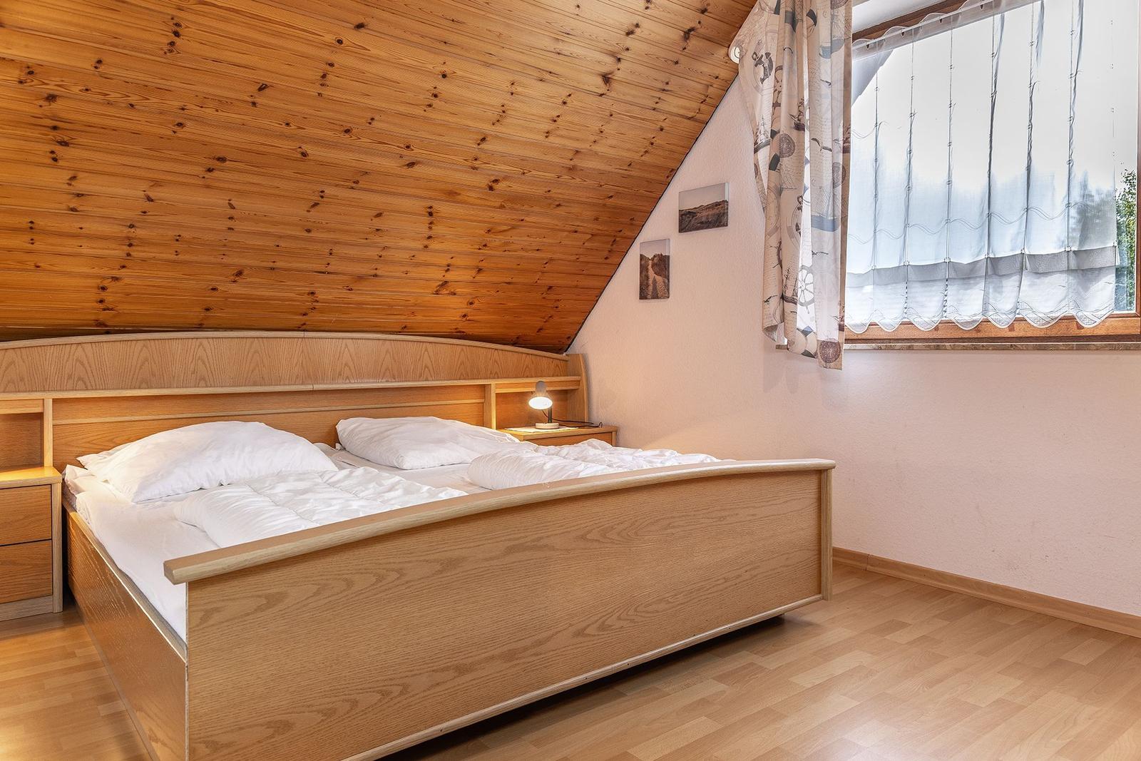Schlafzimmer mit Holzbett, Dachschräge und Fenster mit Vorhängen.