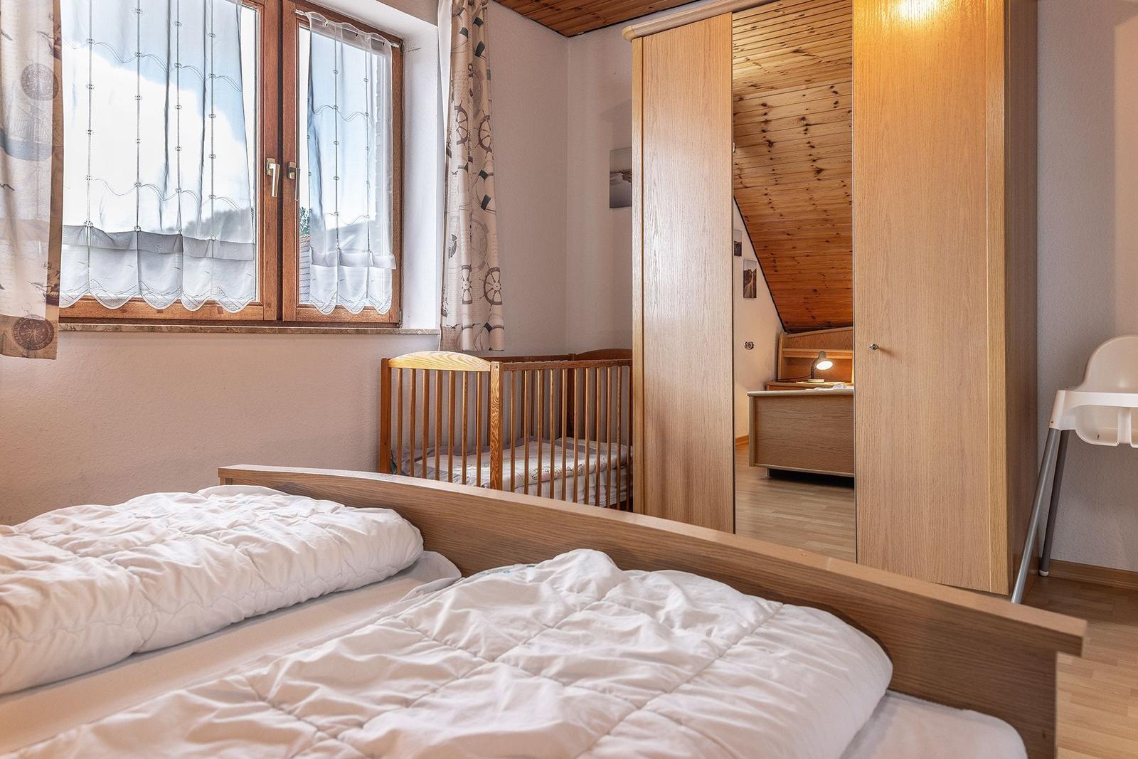 Zimmer mit Doppelbett, Kinderbett und Kleiderschrank. Holzdecke und Fenster mit Vorhängen.