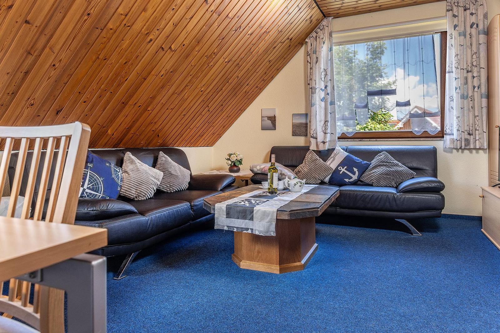 Wohnzimmer mit zwei Sofas, Holzdecke und blauem Teppich. Fenster mit Vorhängen.