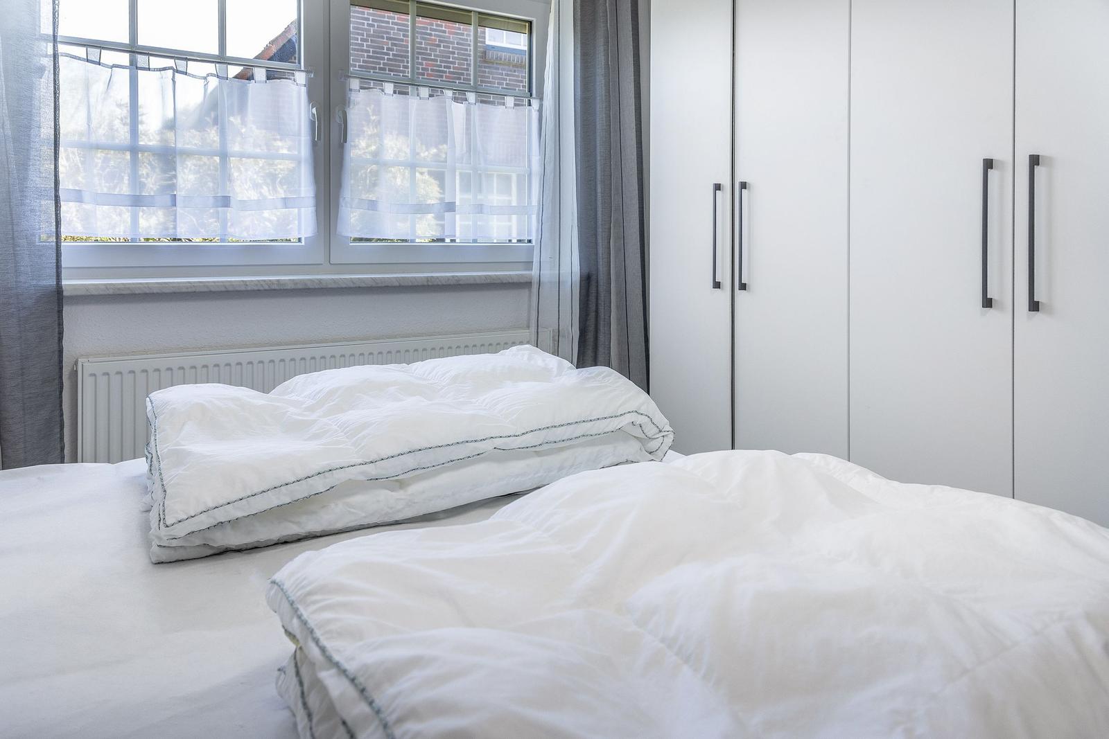 Schlafzimmer mit weißem Bett, Fenster mit Vorhängen und weißen Schränken.