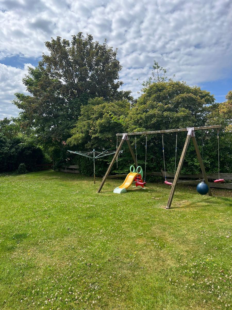 Großer Garten mit Spielplatz, Schaukel und Rutsche. Grasfläche und Bäume im Hintergrund.