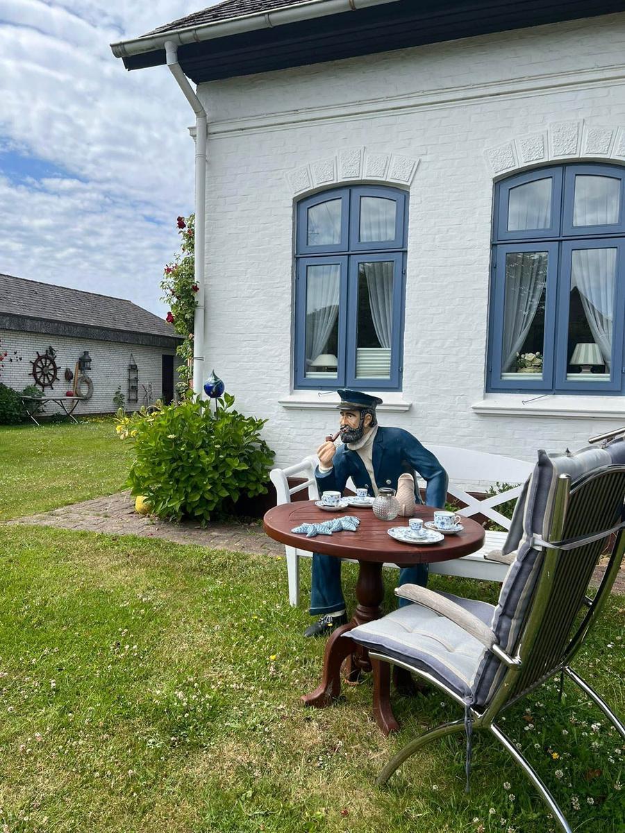 Haus mit weißer Fassade und blauen Fenstern. Auf dem Grundstück steht eine Figur am Tisch.