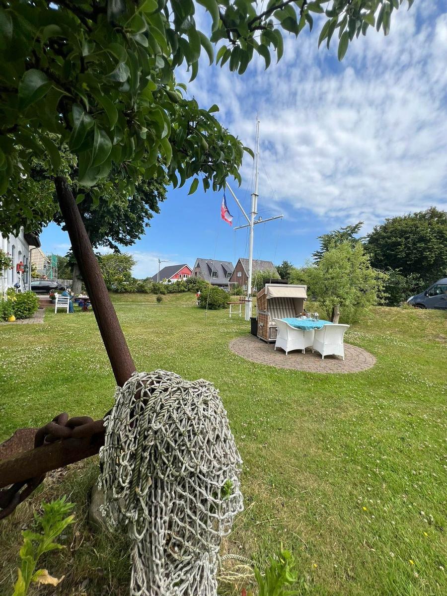 Grüner Garten mit Tisch, Stühlen und Segelboot-Flagge. Vordergrund: großer Anker mit Kette.