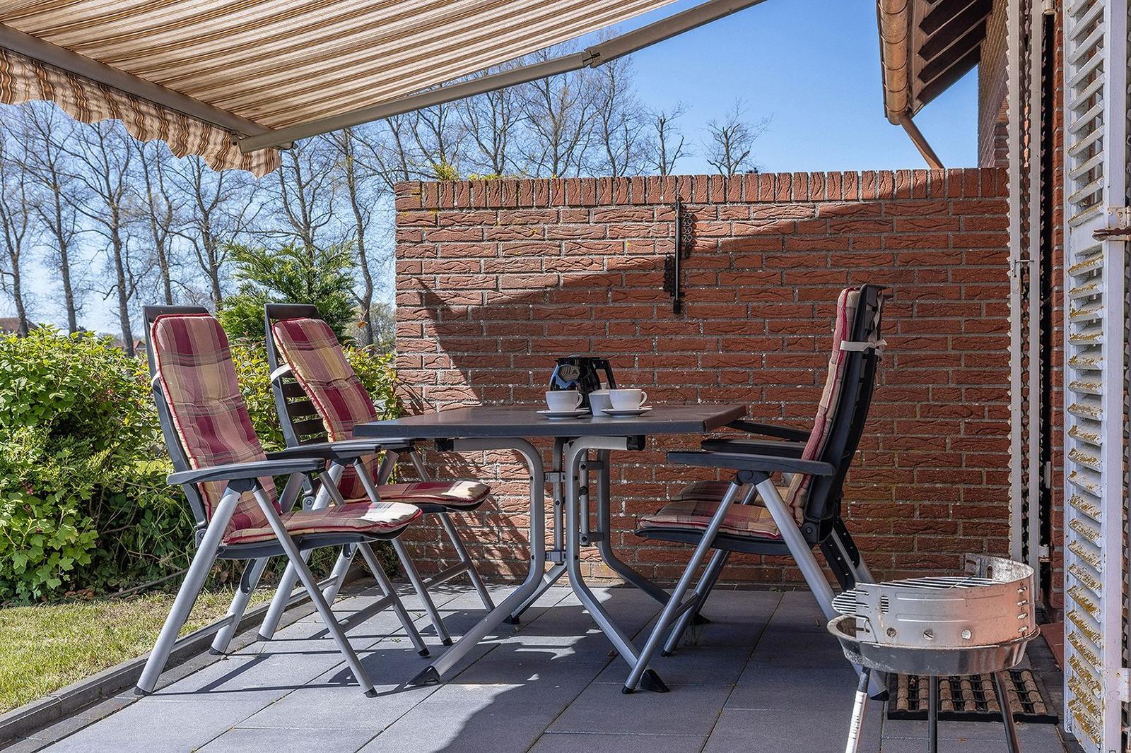 Terrasse mit Tisch, drei Stühlen und Grill unter Markise. Hintergrund: Backsteinmauer und Bäume.