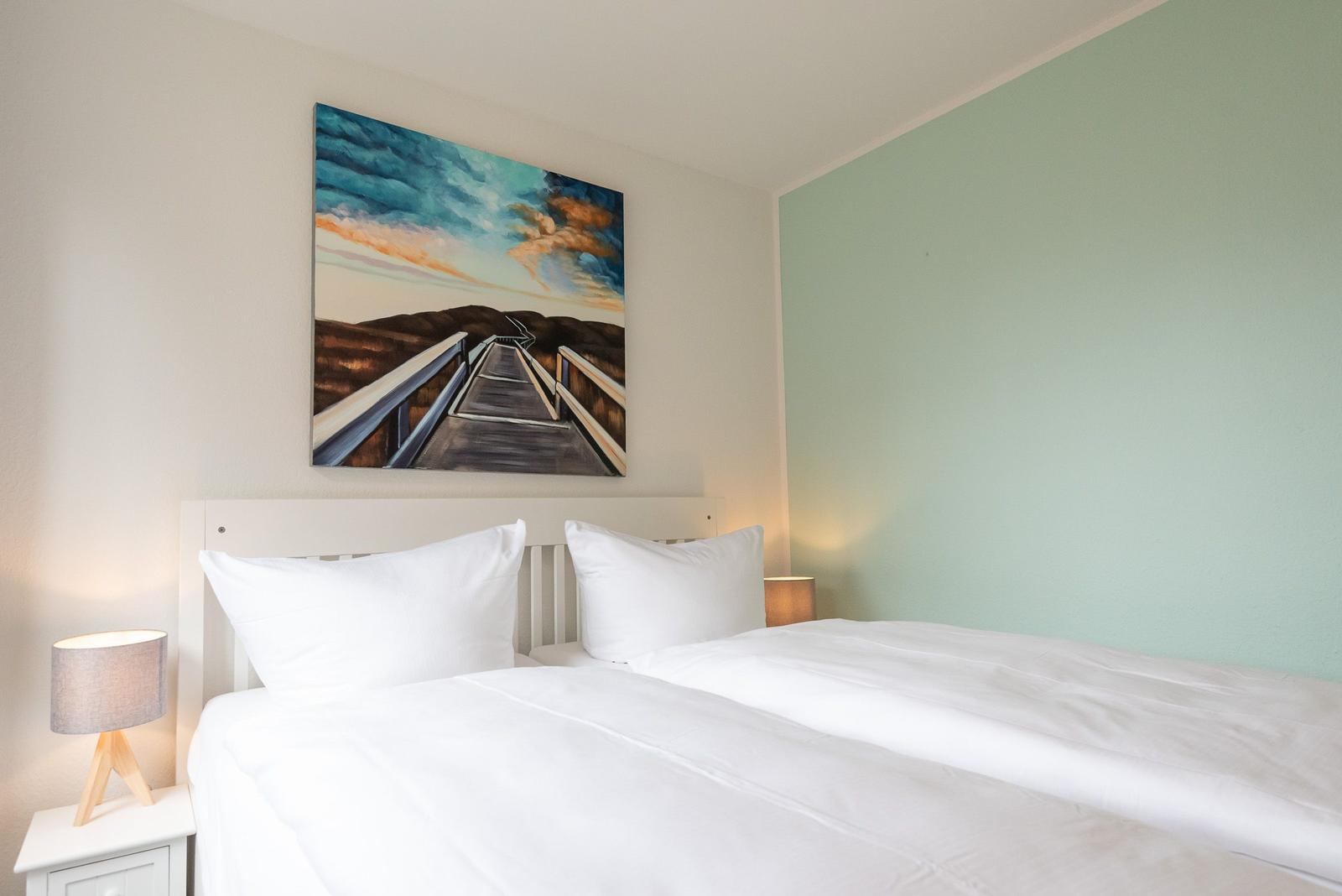 Schlafzimmer mit weißem Bett, Wandbild und Nachttisch mit Lampe.