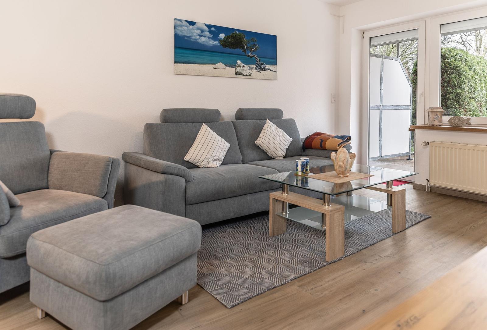 Wohnzimmer mit grauem Sofa, Glas-Tisch und Wandbild. Durchgang zur Terrasse.