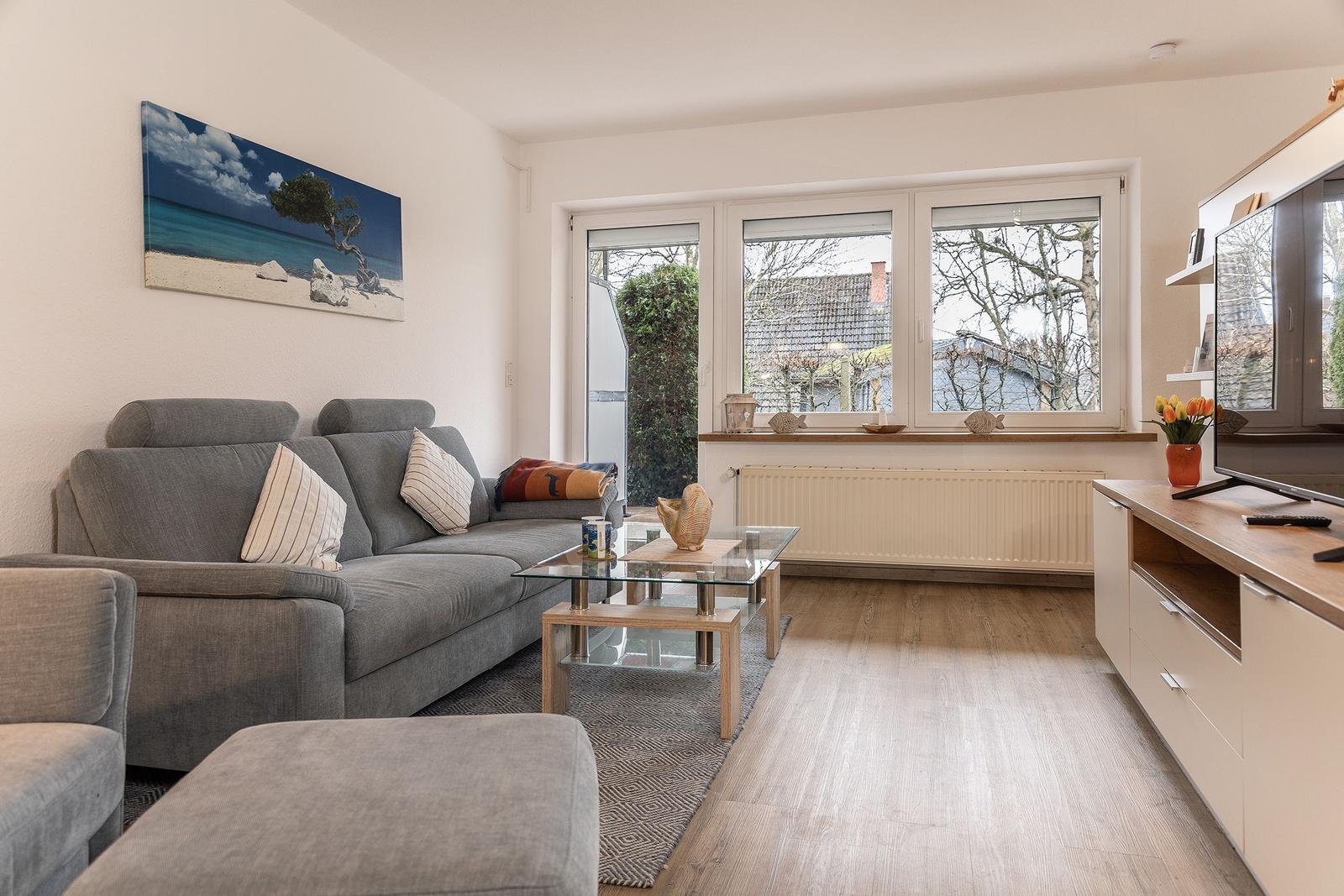 Wohnzimmer mit Sofa, Glas-Tisch und TV-Möbel. Fenster blicken auf Garten und Dächer.