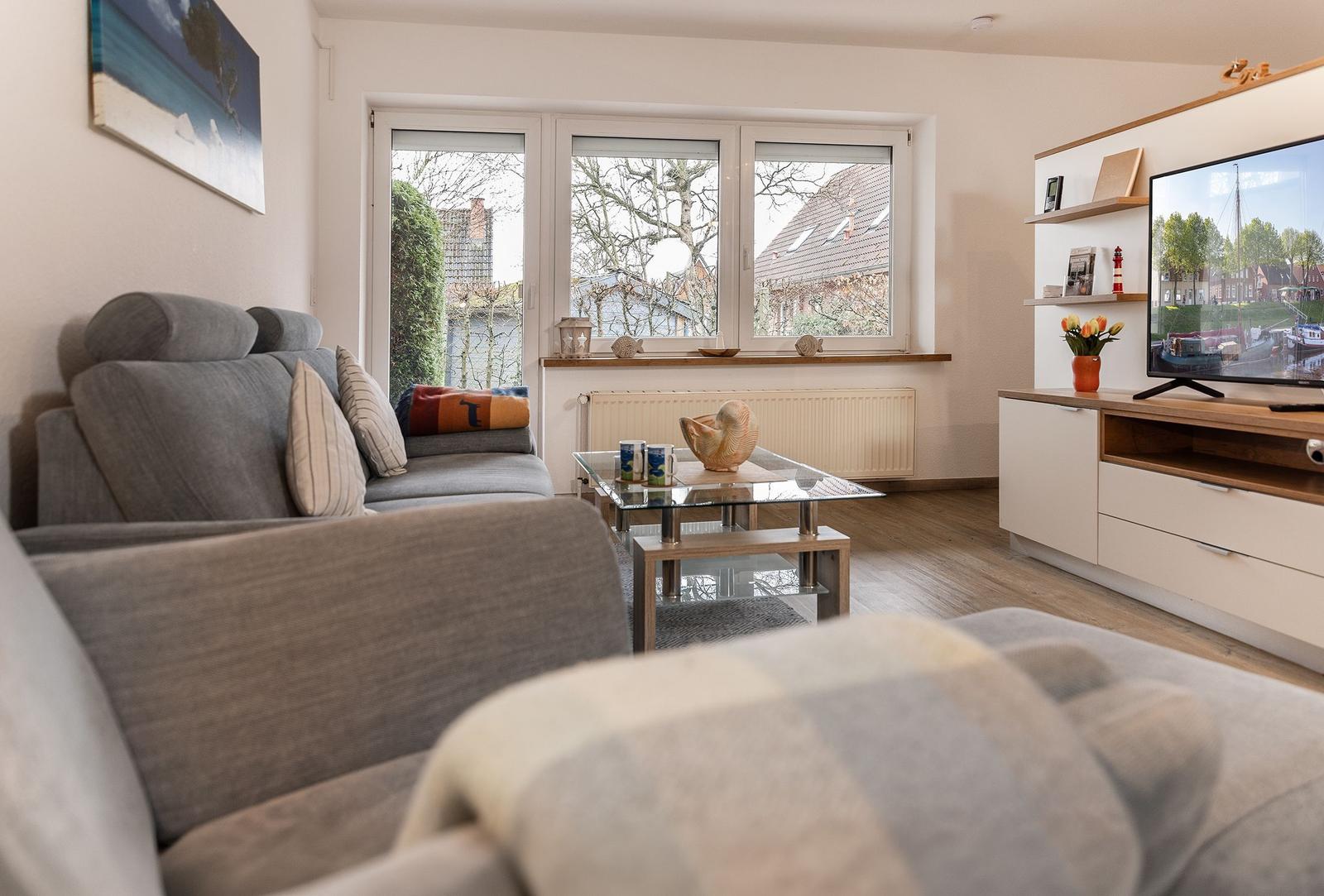Wohnzimmer mit Sofa, Glas-Tisch und Fernseher. Fenster zeigen Außenbereich.